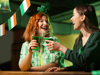 Red-haired pretty girl in a green checkered shirt and a girl with a mask drinking green beer