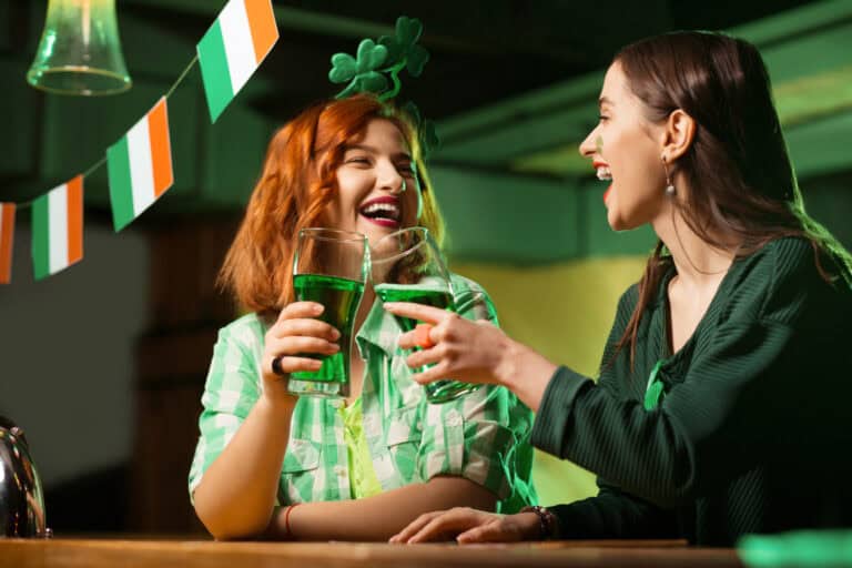 Red-haired pretty girl in a green checkered shirt and a girl with a mask drinking green beer